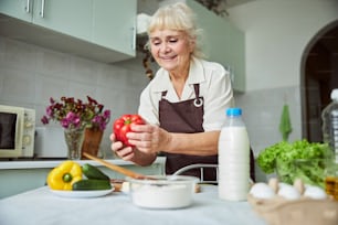 Cheerful elderly lady in apron holding red bell pepper and smiling while standing by the table with vegetables, eggs and milk