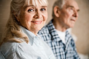 Beaming lady. Pleasant beaming senior lady sitting at the bed with her husband. Men is blurred on the background