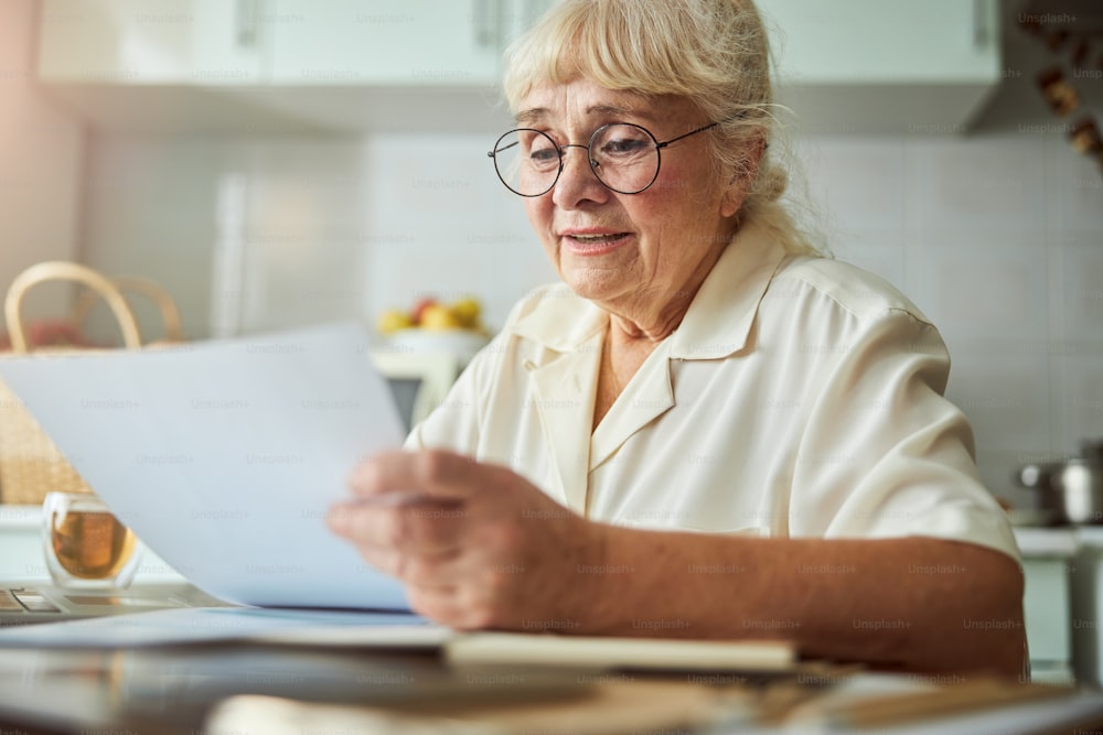 Lovely elderly lady in glasses sitting at the table with cup of tea and holding sheet of paper