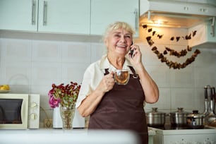 Joyful elderly woman in apron holding cup of tea and smiling while talking on mobile phone at home