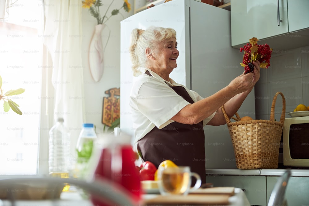 Joyeuse dame âgée en tablier regardant les baies rouges et souriant tout en passant du temps dans la cuisine à la maison