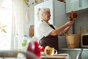 Cheerful senior lady in apron looking at red berries and smiling while spending time in kitchen at home
