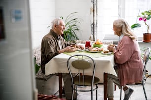 Joyful mature couple is tasting sweet self-baked buns in kitchen. They are sitting and drinking tea
