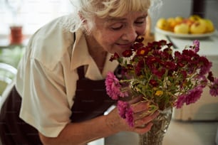 Cheerful elderly lady in apron enjoying the scent of bouquet and smiling