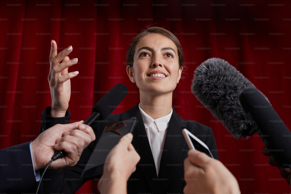 Retrato de cintura para arriba de una funcionaria sonriente dando una entrevista a un periodista y hablando con micrófonos contra la cortina roja