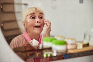 Bella signora anziana che si guarda allo specchio e sorride durante l'utilizzo del prodotto cosmetico per la cura della pelle