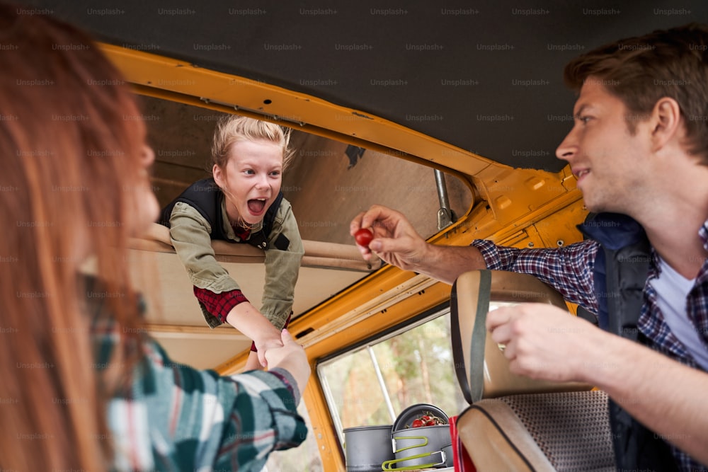 A delighted blondy girl spends time with her parents in motorhome. Daddy feeds his daughter cherry tomato