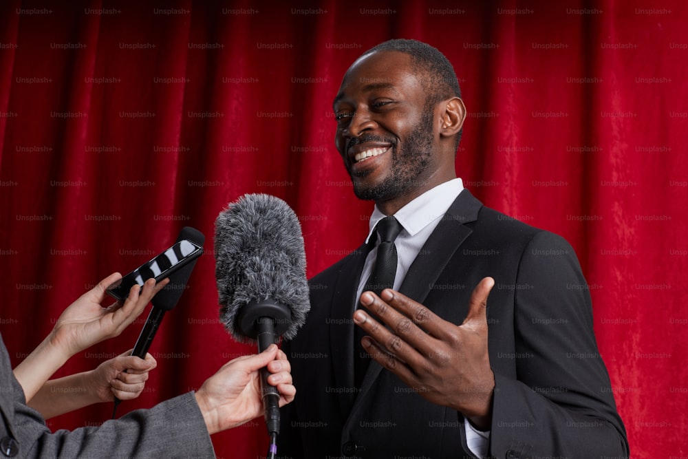 Waist up portrait of smiling African-American man giving interview to journalist and speaking to microphones against red curtain