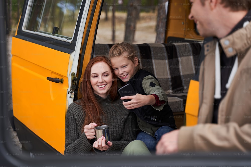 Funny time together. Family drinking tea at the nature while sitting near their car. Cute girl making selfie with her lovely mother. Father admires of his dear people