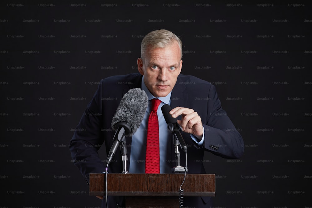 Waist up portrait of mature man standing at podium and speaking to microphone against black background, copy space