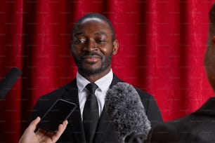 Retrato de homem afro-americano sorridente dando entrevista a jornalista e falando aos microfones contra cortina vermelha
