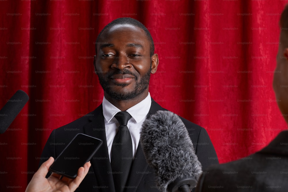 Portrait of smiling African-American man giving interview to journalist and speaking to microphones against red curtain