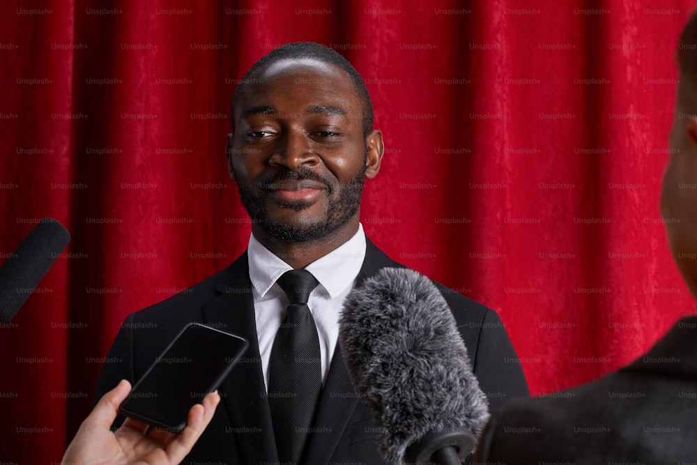 Portrait d’un homme afro-américain souriant donnant une interview à un journaliste et parlant aux micros contre un rideau rouge