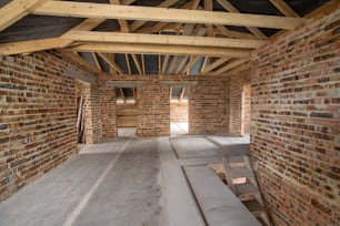 Interior de casa de ladrillo sin terminar con piso de concreto, paredes desnudas listas para enlucido y marco de techo de madera ático en construcción.