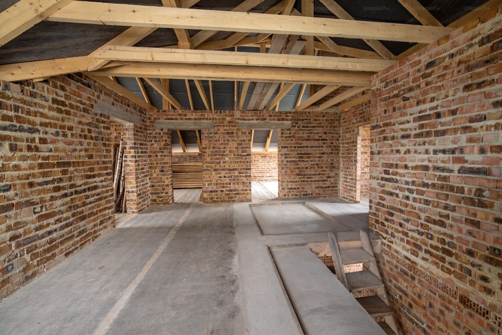 コンクリートの床、左官工事の準備ができている裸の壁、建設中の木製屋根ふきフレーム屋根裏部屋を持つ未完成のレンガ造りの家のインテリア。