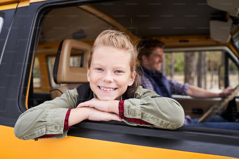 父親と一緒に車で森の中を運転しながら、窓の外を見て良い週末を楽しむ金髪の陽気な女の子