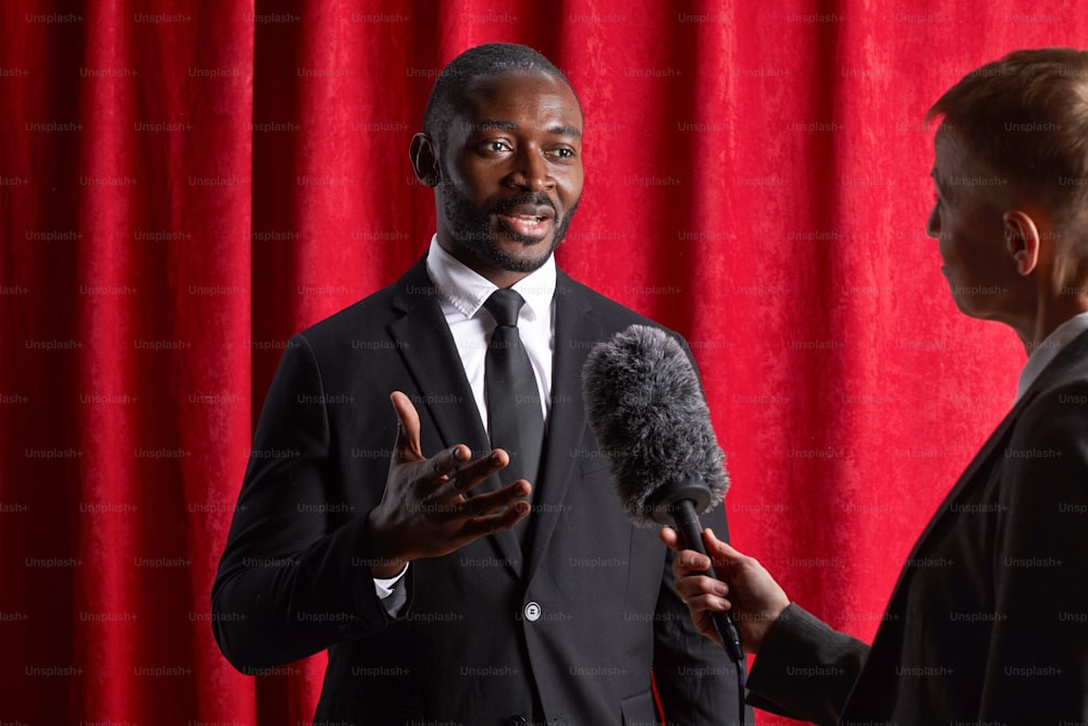 Retrato de homem afro-americano dando entrevista a jornalista e falando aos microfones contra cortina vermelha