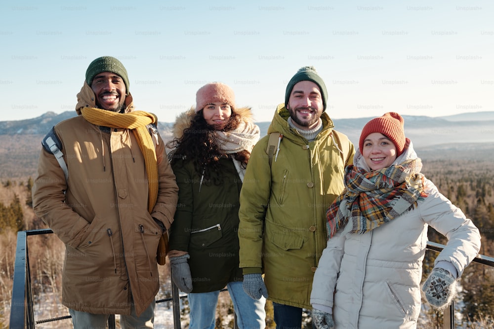 Happy young intercultural friends in winterwear standing in front of camera against blue sky over mountains covered with snow while chilling out