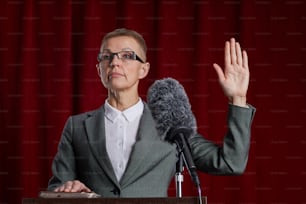 Waist up portrait of mature woman giving oath while standing at podium on stage against red curtain, copy space