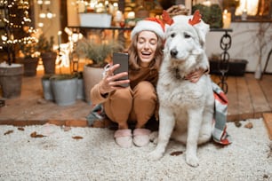Portrait of a woman in christmas hat with her cute dog celebrating a New Year holidays on the beautifully decorated terrace at home, making selfie photo together