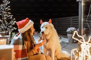 Retrato de una mujer con sombrero navideño y a cuadros con su lindo perro celebrando unas vacaciones de Año Nuevo en la terraza bellamente decorada de su casa, alimentando al perro con galletas de jengibre
