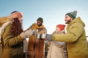 Happy young friends in winterwear cheering up with hot tea and chatting against blue sky over mountains and forest on winter weekend