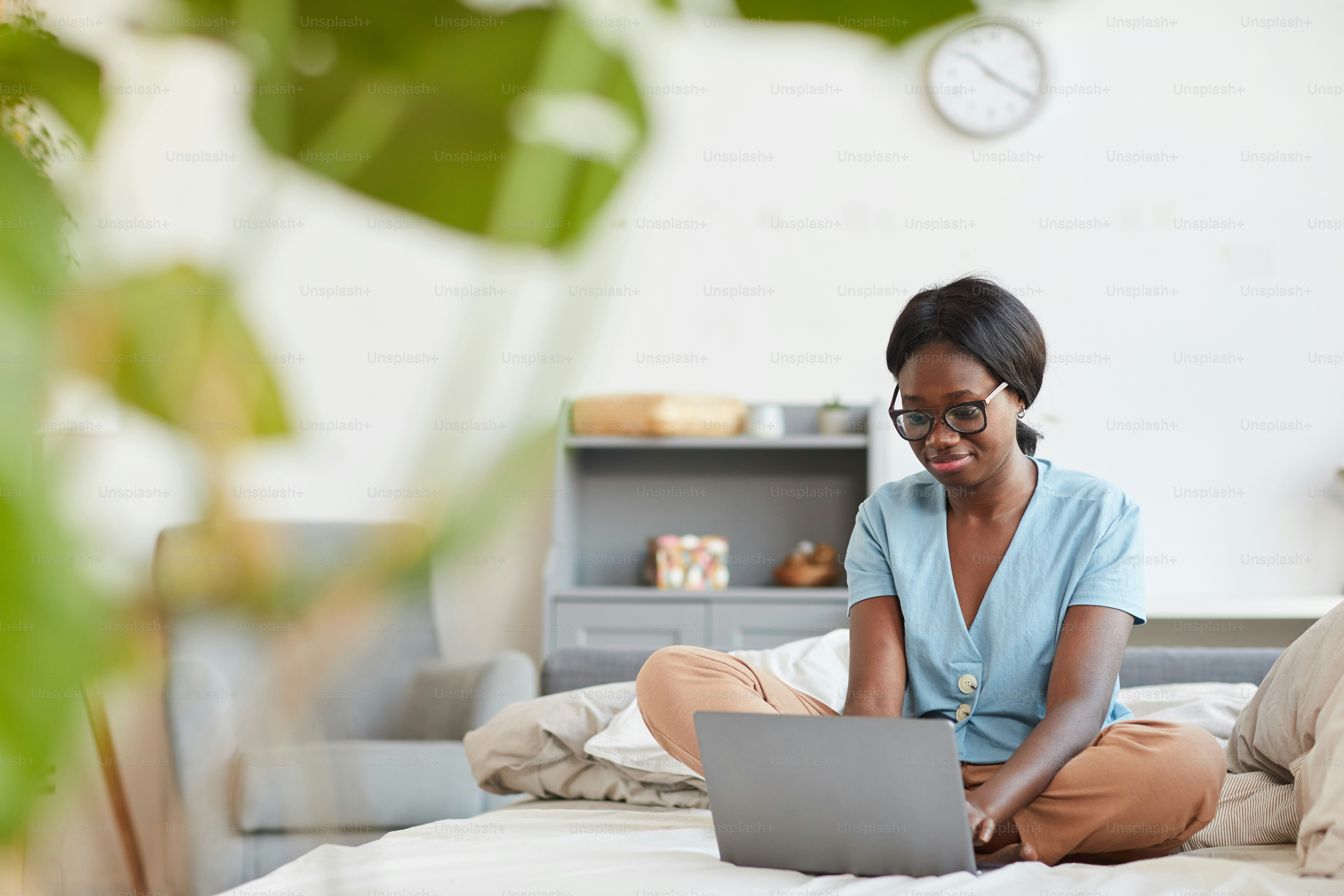 Potret panjang penuh wanita muda Afrika-Amerika menggunakan laptop sambil duduk di tempat tidur di rumah dengan bingkai daun tanaman, ruang salinan