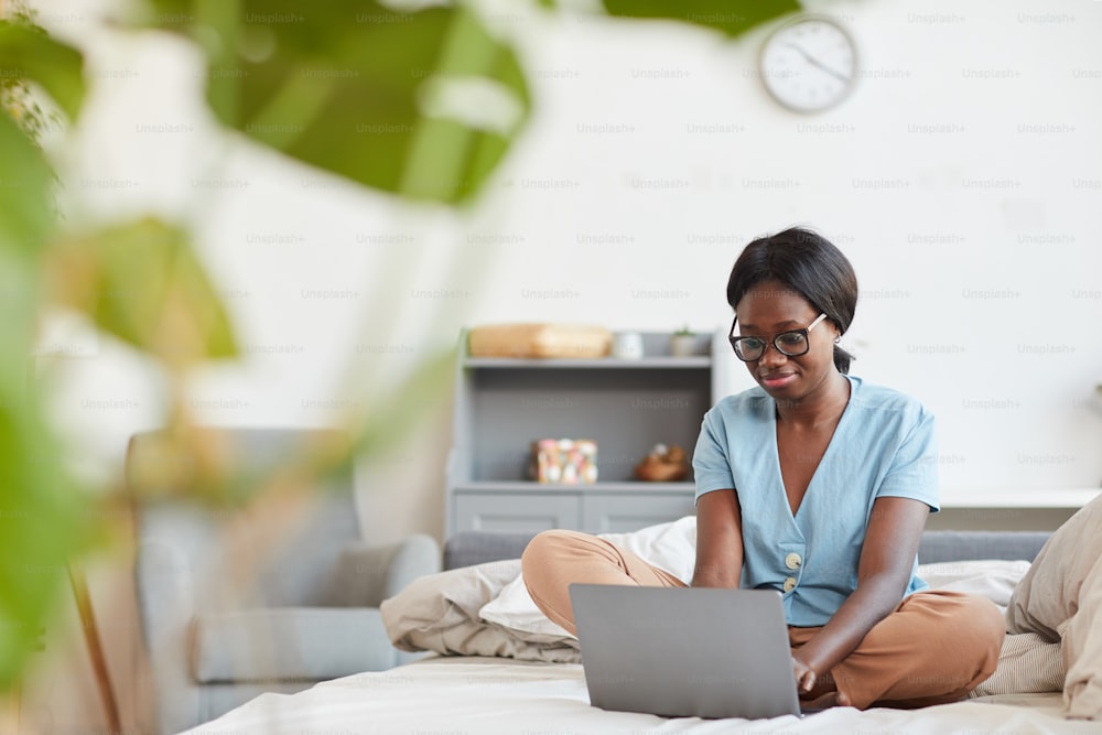Ritratto a figura intera di giovane donna afroamericana che utilizza il computer portatile mentre è seduta sul letto a casa con l'inquadratura delle foglie delle piante, lo spazio di copia