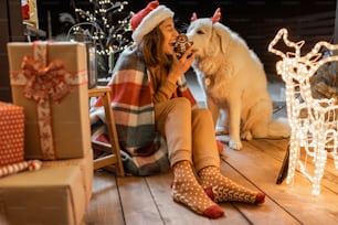 Ritratto di una donna in cappello di Natale e plaid con il suo simpatico cane che festeggia le vacanze di Capodanno sulla terrazza splendidamente decorata a casa, nutrendo il cane con biscotti di pan di zenzero