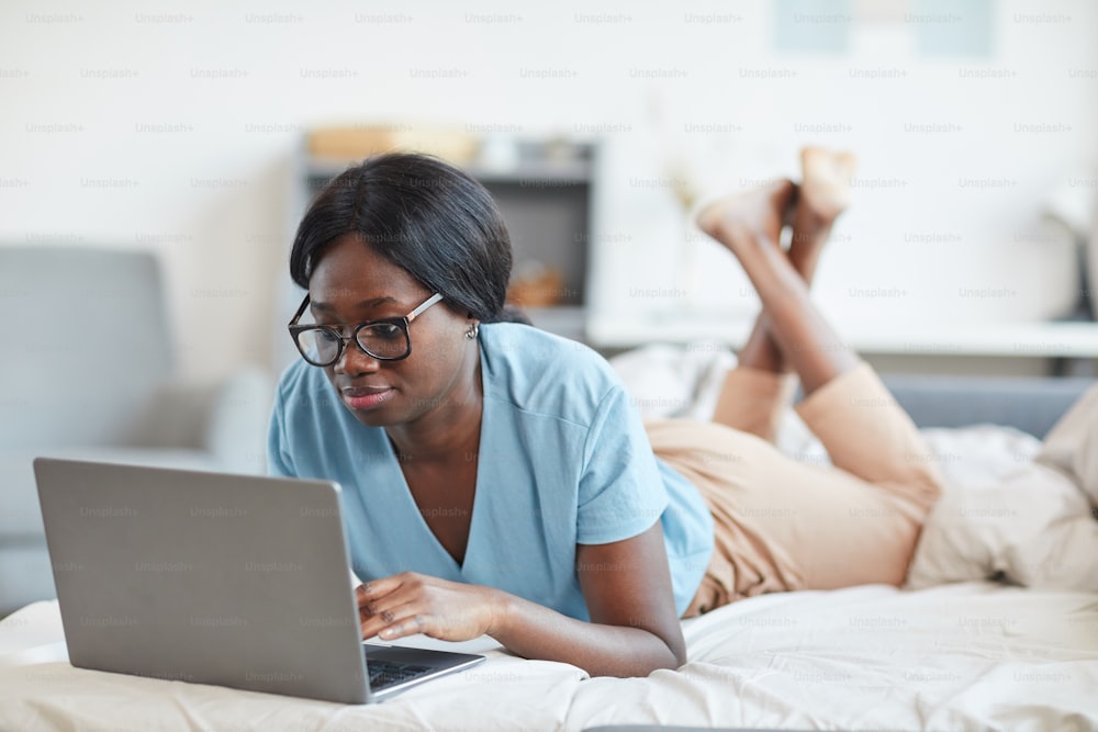 Ritratto di giovane donna afroamericana che utilizza il computer portatile mentre è sdraiata a letto e si gode il fine settimana a casa con comodità, spazio di copia