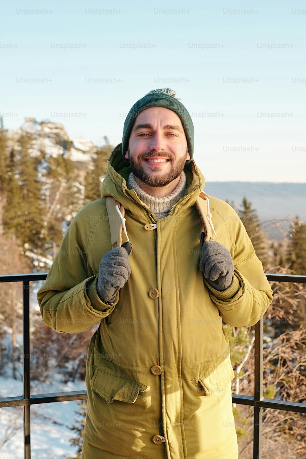 Young smiling man with backpack standing in front of camera against trees and rocks covered with snow and clear blue sky above on winter day