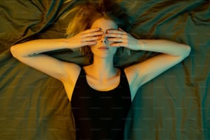 View of young blond relaxed female in black leotard covering eyes with hands while lying on bed in the morning reluctant to wake up