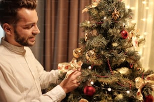 白いシャツを着た幸せな若いひげを生やした男性が、クリスマスツリーに装飾的な絹の弓を置き、次のお祝いのために家を準備する