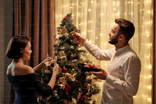 Joven morena con elegante vestido negro y su marido barbudo poniendo decoraciones en el árbol de Navidad mientras se preparan para la celebración