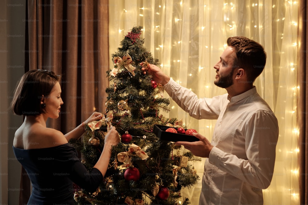 Young brunette woman in elegant black dress and her bearded husband putting decorations on Christmas tree while preparing for celebration