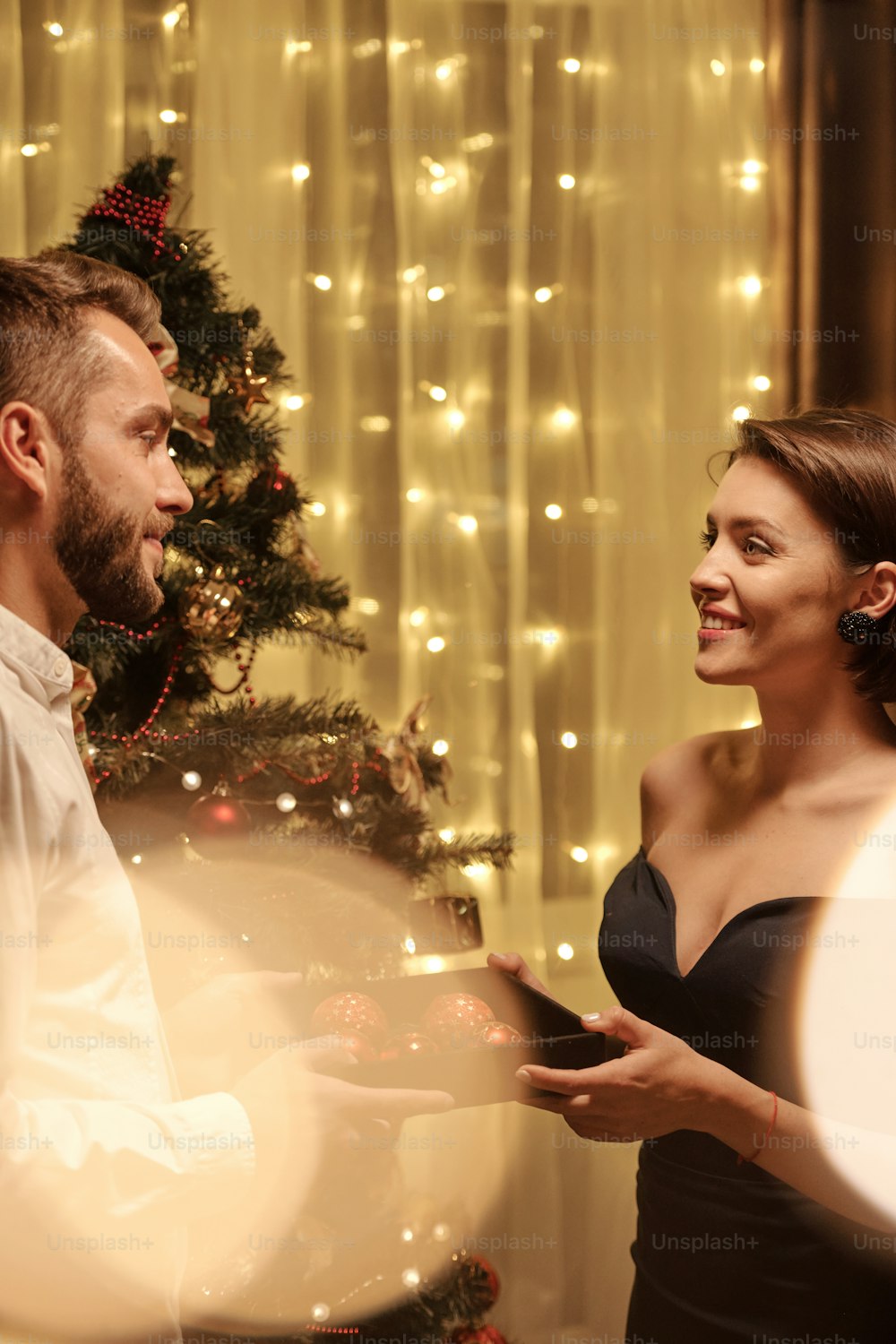 Happy elegant young woman giving red toy balls to her husband while looking at him with smile against Christmas tree in home environemnt