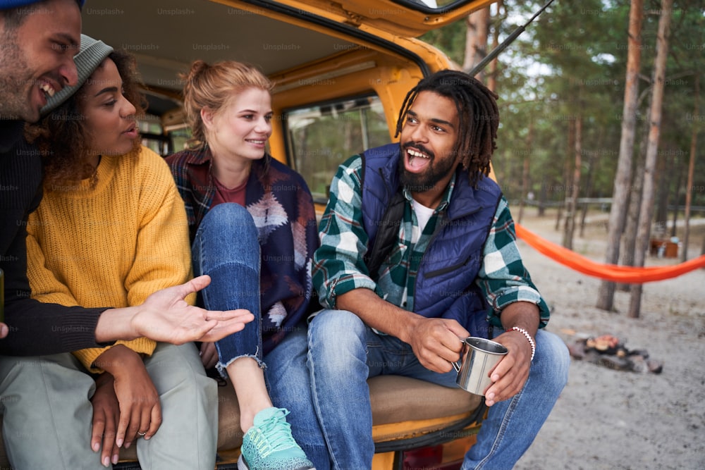Spending time together. Portrait of multiracial young people sitting at the car trunk drinking tea and telling funny stories