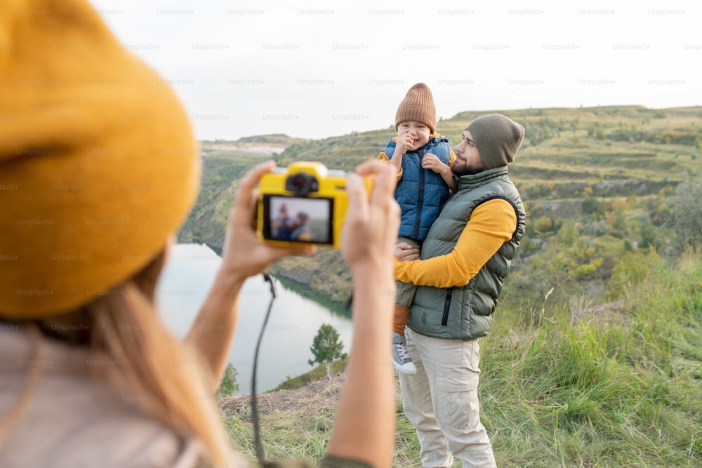 Young female with photocamera taking photograph of her husband and little son standing against lake surrounded by mountains during trip