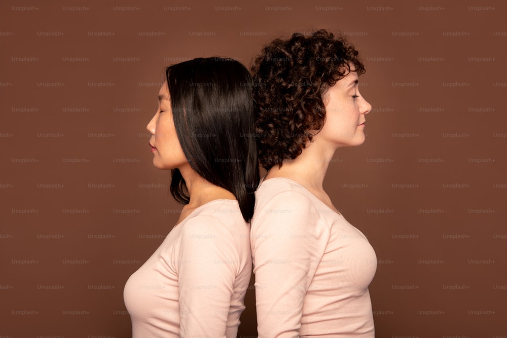 Two young intercultural women in white pullovers keeping eyes closed while standing close to one another on brown background