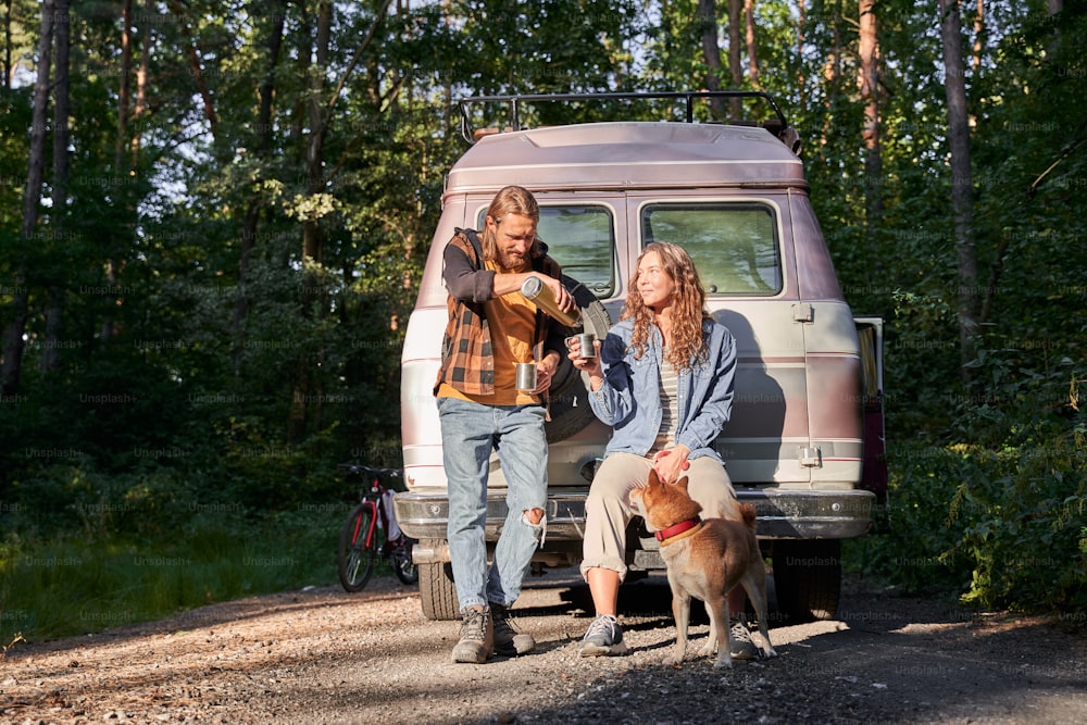 Young couple travelers in casual outfits travelling with their dog. Man pours tea from thermos for his girlfriend. Local tourism, weekend trip