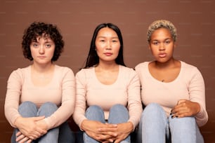 Group of contemporary young serious females of various ethnicities sitting in front of camera close to one another against brown background