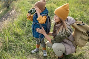 Junge Backpackerin in warmer Freizeitkleidung hockt bei ihrem entzückenden kleinen Sohn mit Fernglas während eines Wochenendausflugs in natürlicher Umgebung