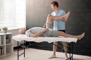 Male physiotherapist holding patient leg bent in knee while helping him with one of physical exercises in rehabilitation center or clinics
