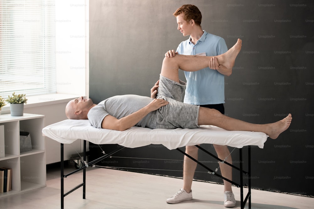 Fisioterapeuta do sexo masculino segurando a perna do paciente dobrada no joelho enquanto o ajuda com um dos exercícios físicos em centro de reabilitação ou clínicas