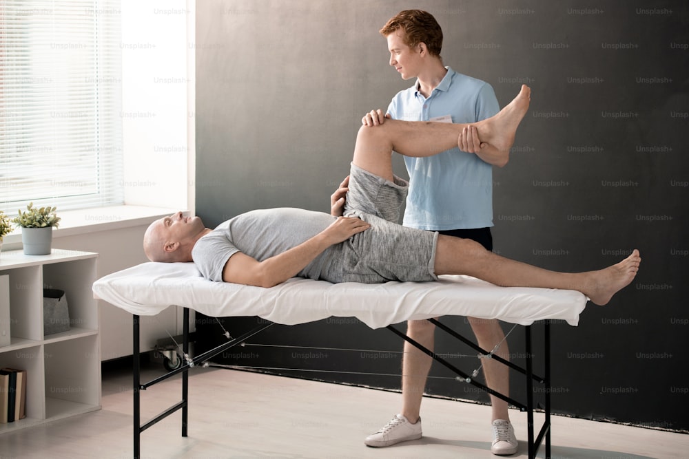 Male physiotherapist holding patient leg bent in knee while helping him with one of physical exercises in rehabilitation center or clinics