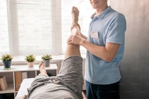 Young physiotherapist holding leg of patient while helping him with one of physical exercises in modern rehabilitation center or clinics