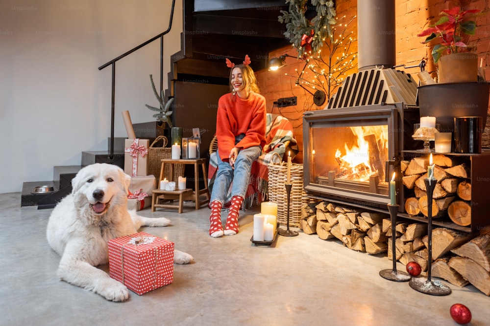 Junge Frau mit ihrem süßen weißen Hund packt Geschenkboxen am Kamin während eines glücklichen Neujahrsurlaubs zu Hause aus