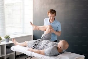 Fisioterapeuta masculino serio mirando a su paciente con la pierna doblada en la rodilla mientras lo ayuda con uno de los ejercicios físicos en el centro de rehabilitación