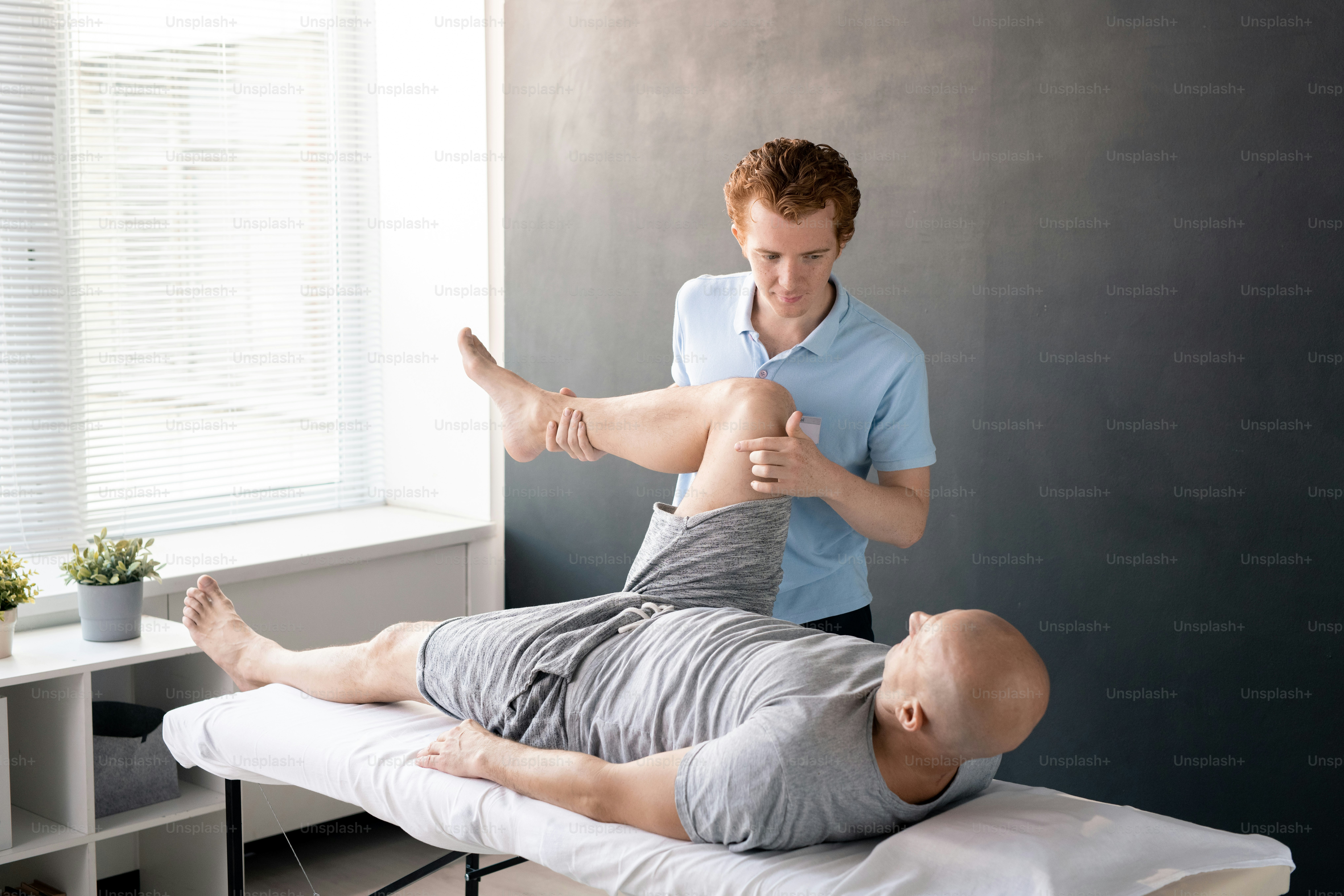 Serious male physiotherapist looking at his patient with leg bent in knee while helping him with one of physical exercises in rehabilitation center