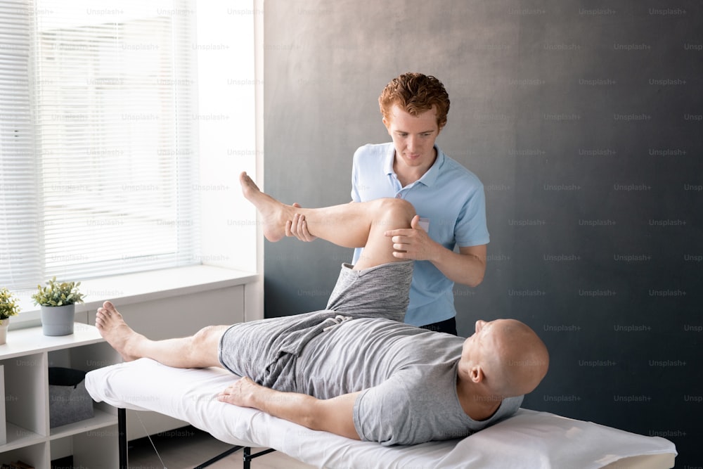 Serious male physiotherapist looking at his patient with leg bent in knee while helping him with one of physical exercises in rehabilitation center
